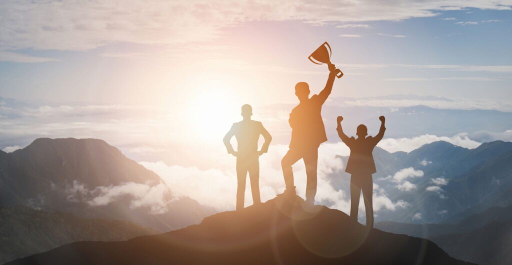 Drei Silhouetten auf einem Berggipfel bei Sonnenaufgang, die mittlere hält einen Pokal hoch - Erfolgs-Projekt im Gesundheitswesen
