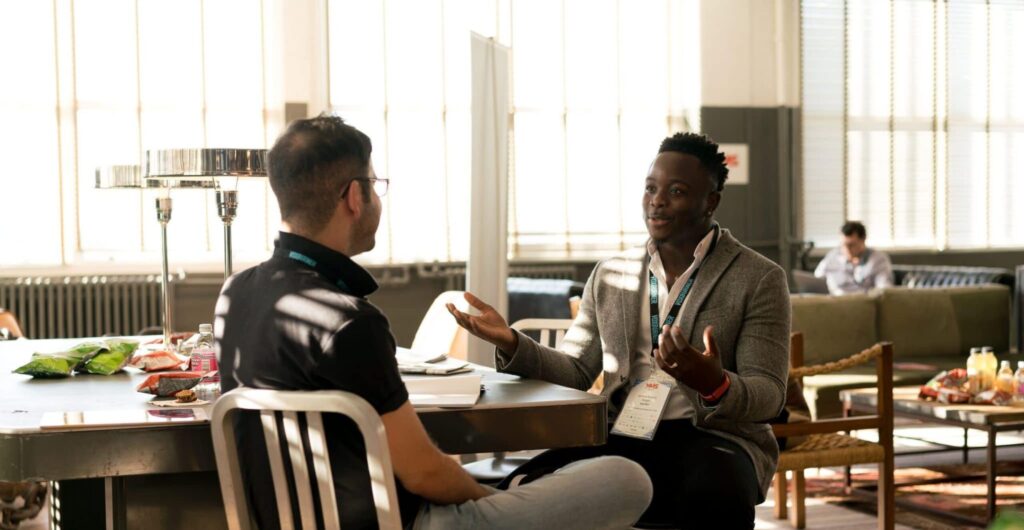 Zwei Kollegen mit Konferenz-Badges im lockeren Gespräch an einem Tisch - Insight zur Marketingberatung